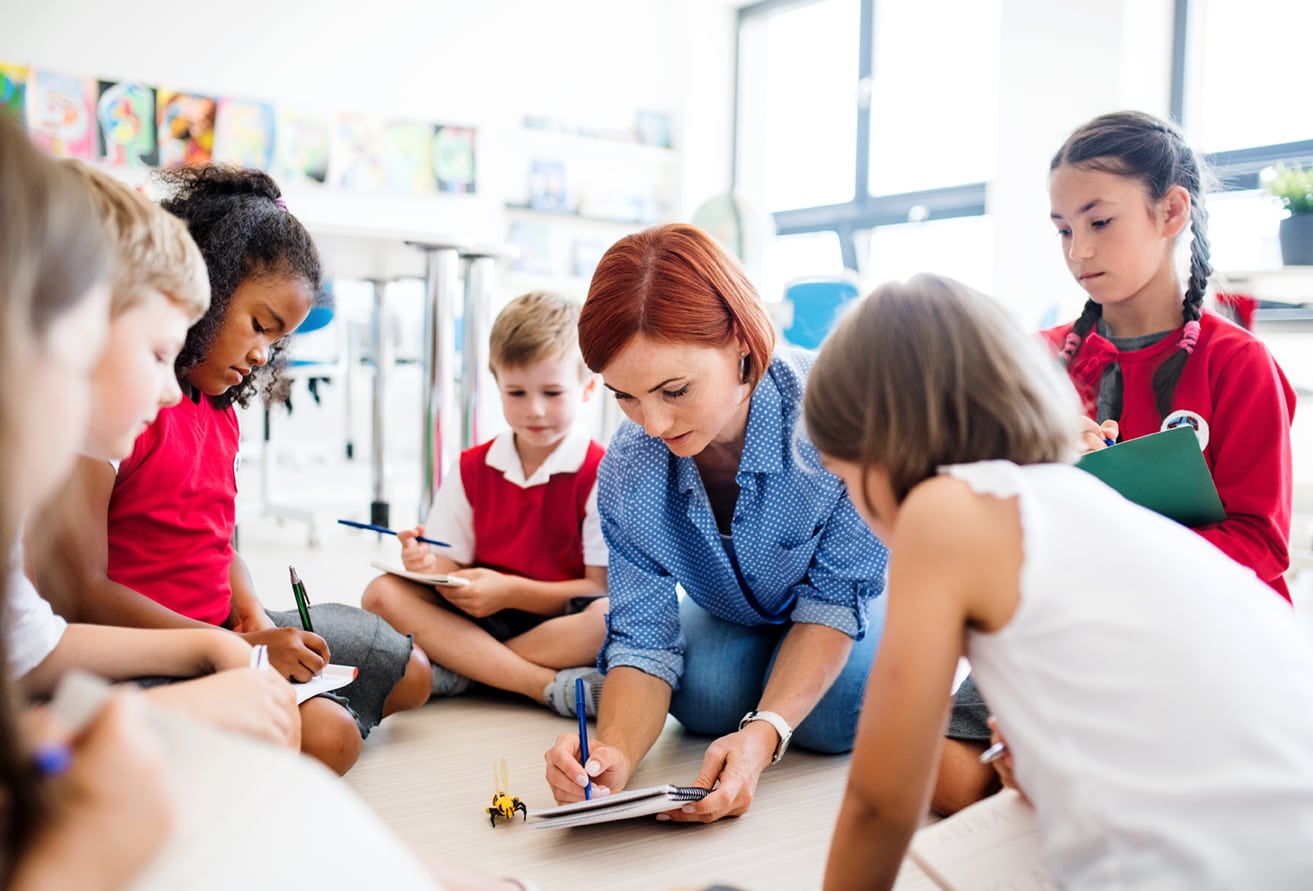 Small schools. Дети и педагог сидят на полу. Педагог сидит 6 на полу. Дети сидят учитель показывает палочкой.